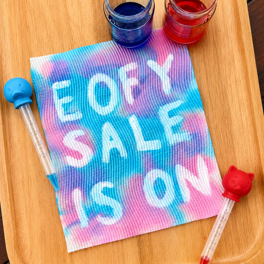 Colorful towel with 'End of Year Sale is On' text on a wooden tray with two small containers of blue and red liquid.